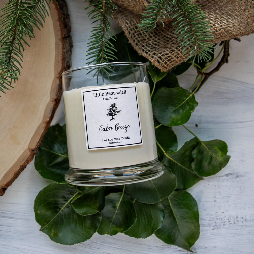 A white candle with a clear glass jar, labeled 'Calm Breeze' by Linda Breeze, placed on a white surface with green foliage in the background.