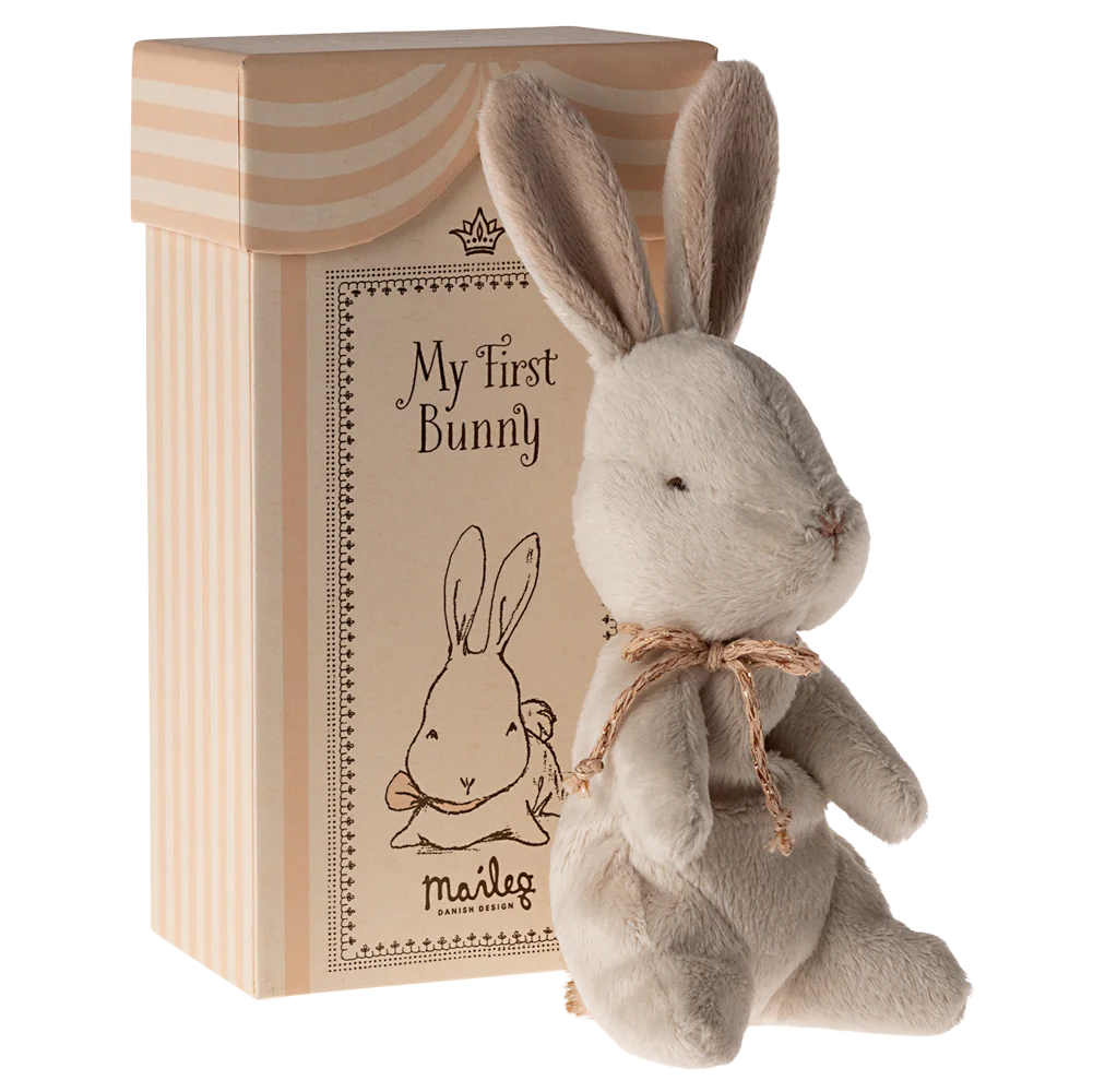 A soft plush toy rabbit with an off-white color, sitting next to its beige cardboard gift box which has 'My First Bunny' written on it.