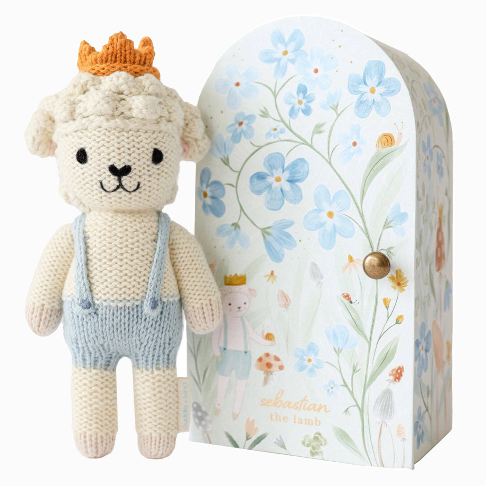 Knitted toy sheep with a crown and overalls next to a floral-patterned box
