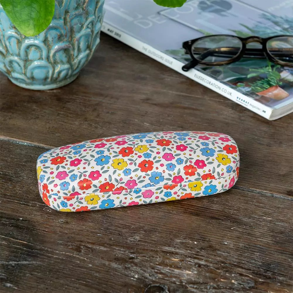 Floral-patterned glasses case on a wooden surface with a book and plant in the background