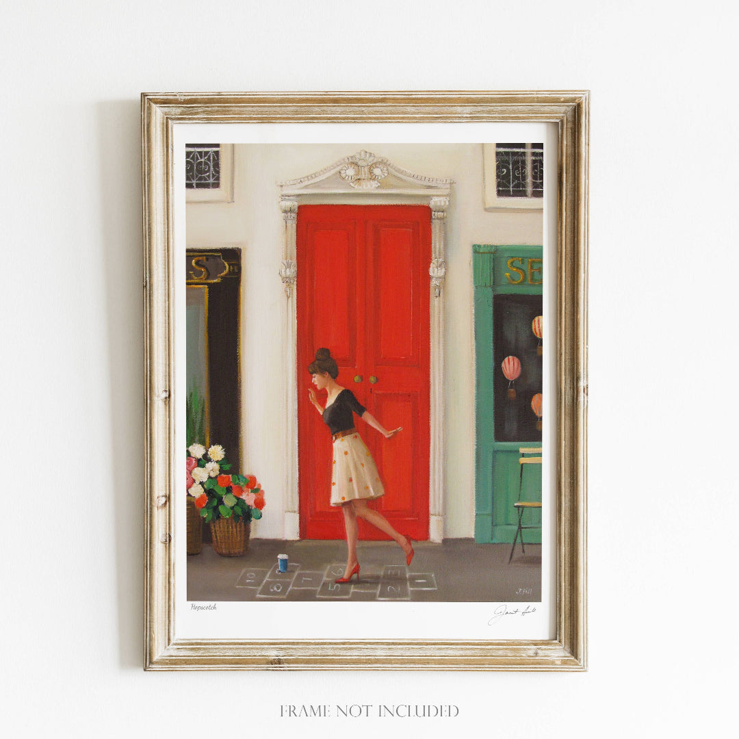 A framed art print featuring a vibrant scene with a woman playing hopscotch on a red door, with an arched doorway and a decorative flower pot visible.