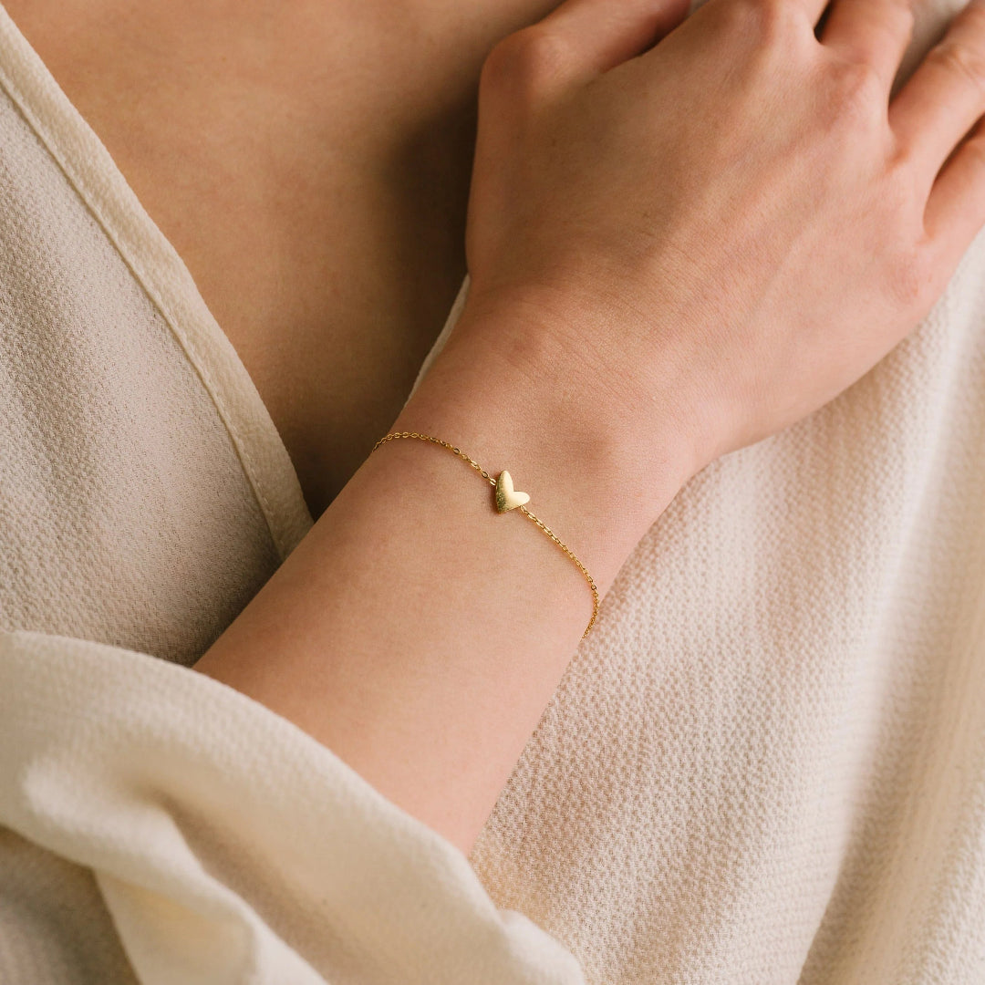 Gold bracelet with a heart charm on a person's wrist against a neutral background