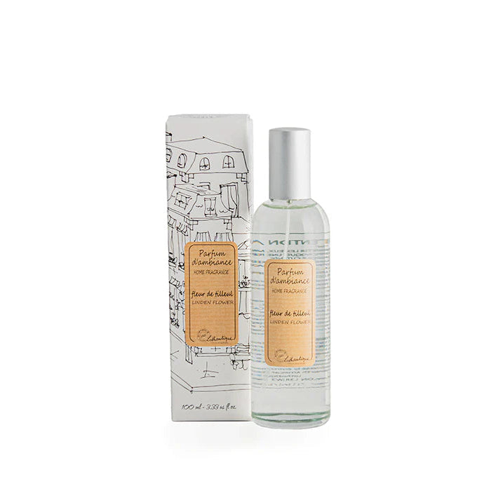 A clear glass room spray bottle with a label next to its packaging featuring a drawing of a house.