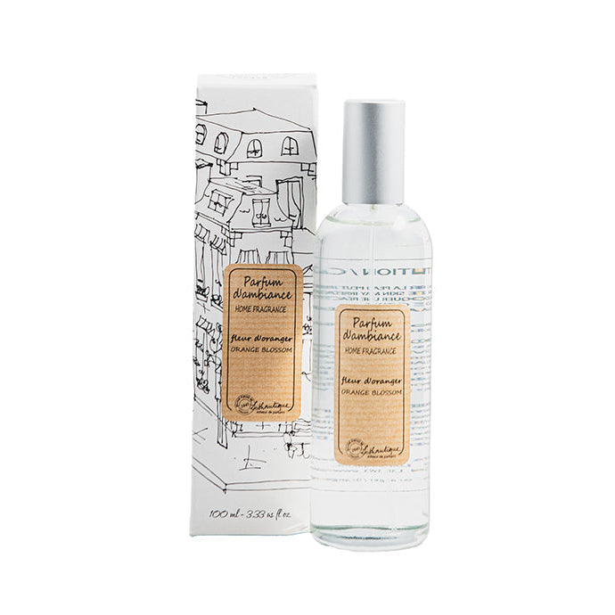 A clear glass room spray bottle with a label next to its packaging featuring a drawing of a house.