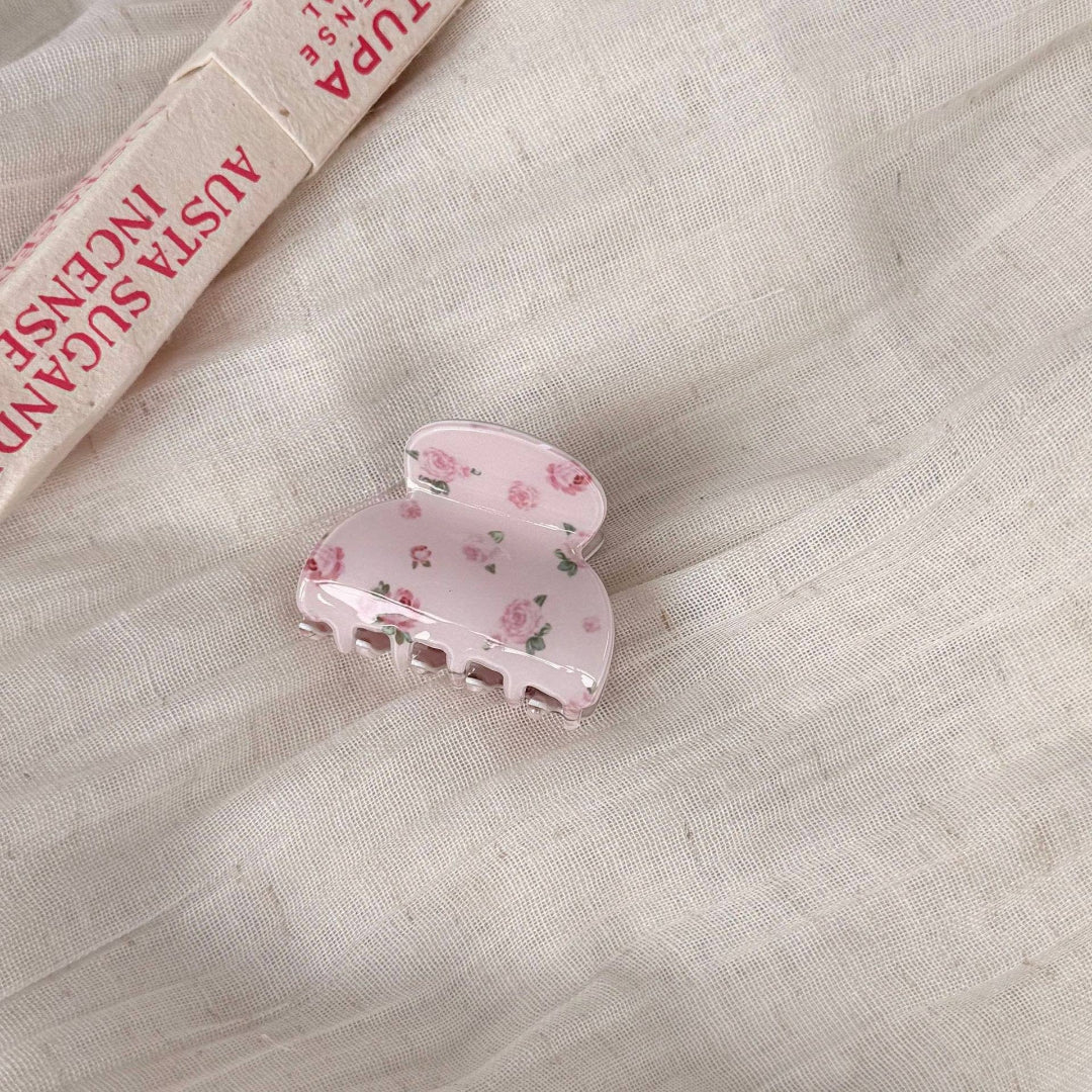 A light pink hair claw clip with a delicate rose pattern, resting on a textured beige linen fabric