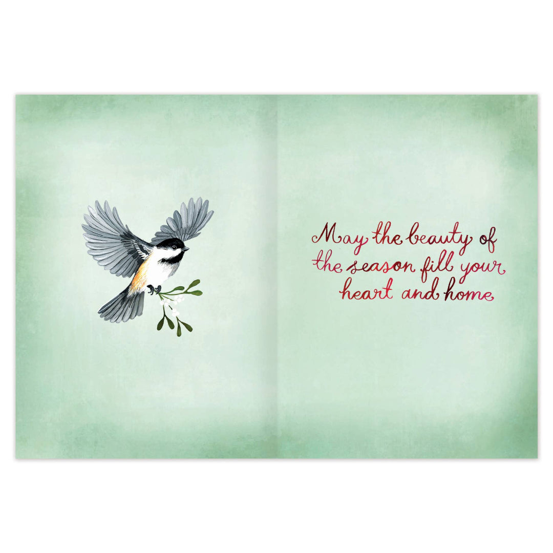 An open greeting card with a pale mint-green watercolor background. The left panel shows a small bird in flight holding a green branch, and the right panel has red cursive text reading, 'May the beauty of the season fill your heart and home.
