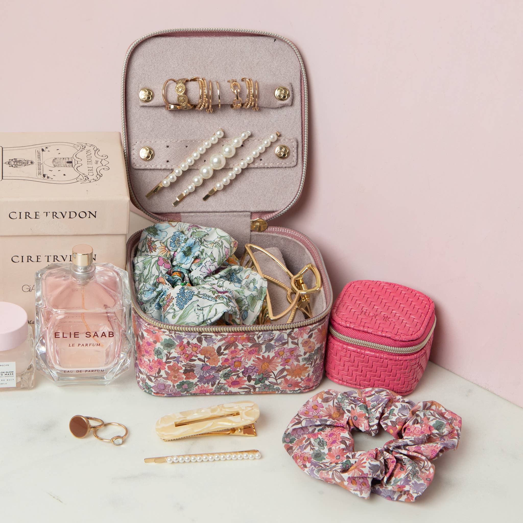 Makeup case with beauty products including scrunchies, hairpins, and perfume on a pink background