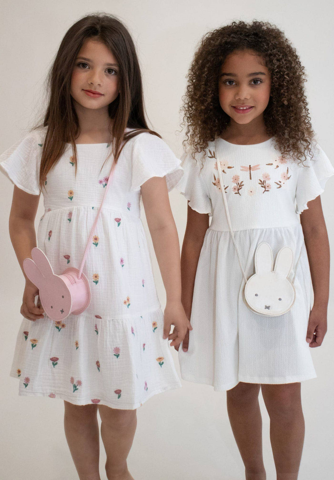 Two young girls wearing white dresses with floral embroidery, holding small bags.