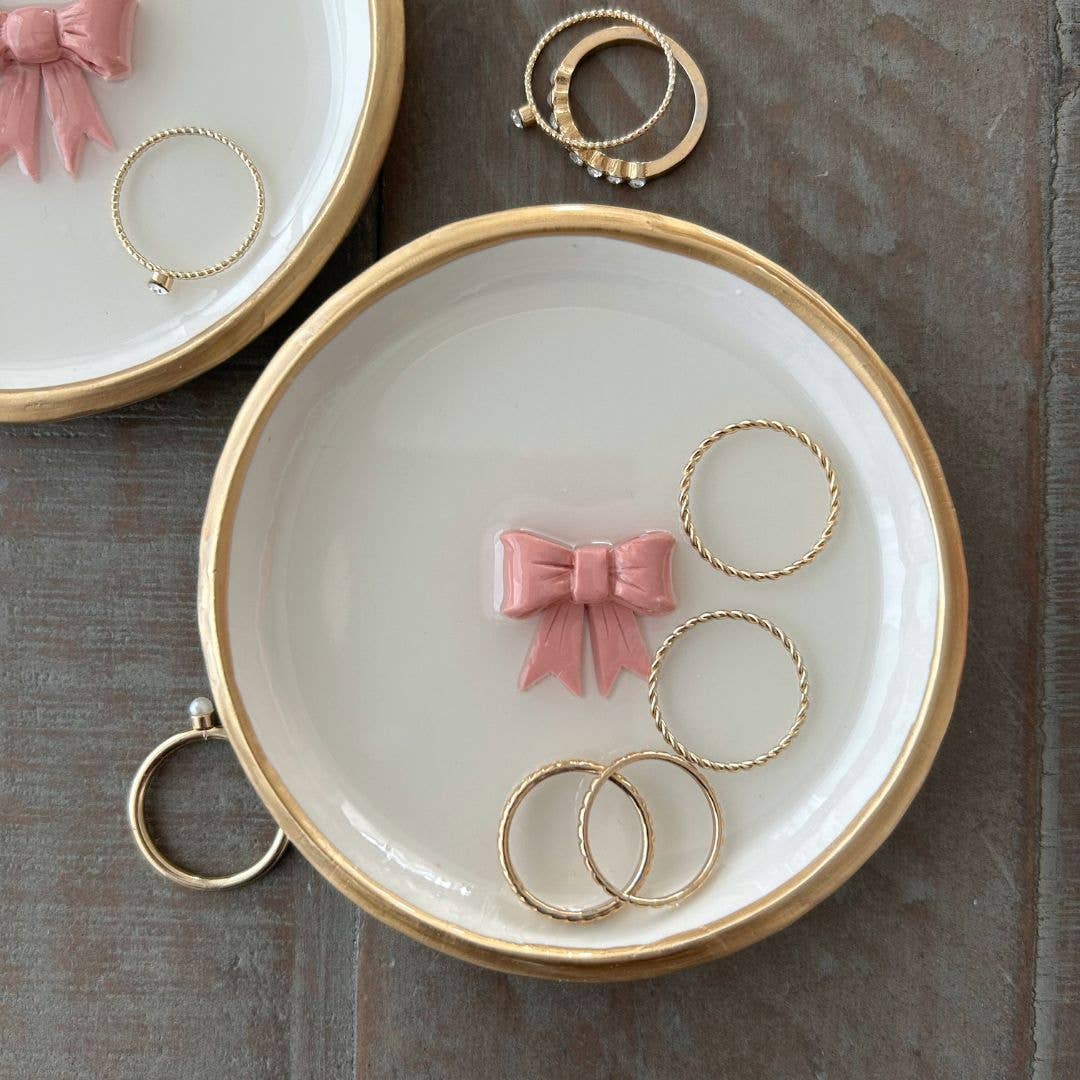 A ring dish with a pink bow design, accompanied by a golden ring, placed on a wooden surface.