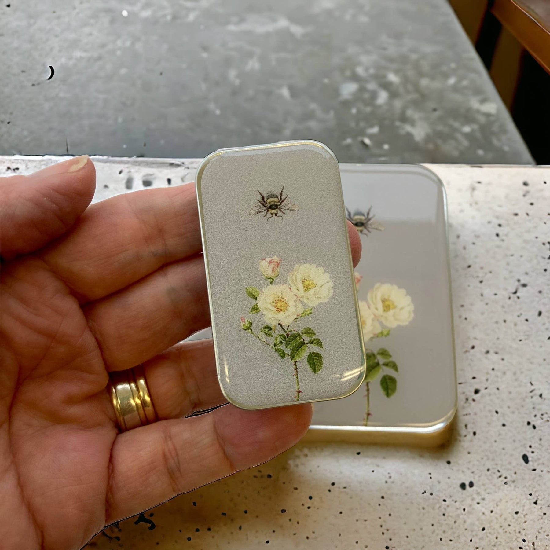 A small rectangular tin box with a vintage rose and bee design, held in a person's hand.