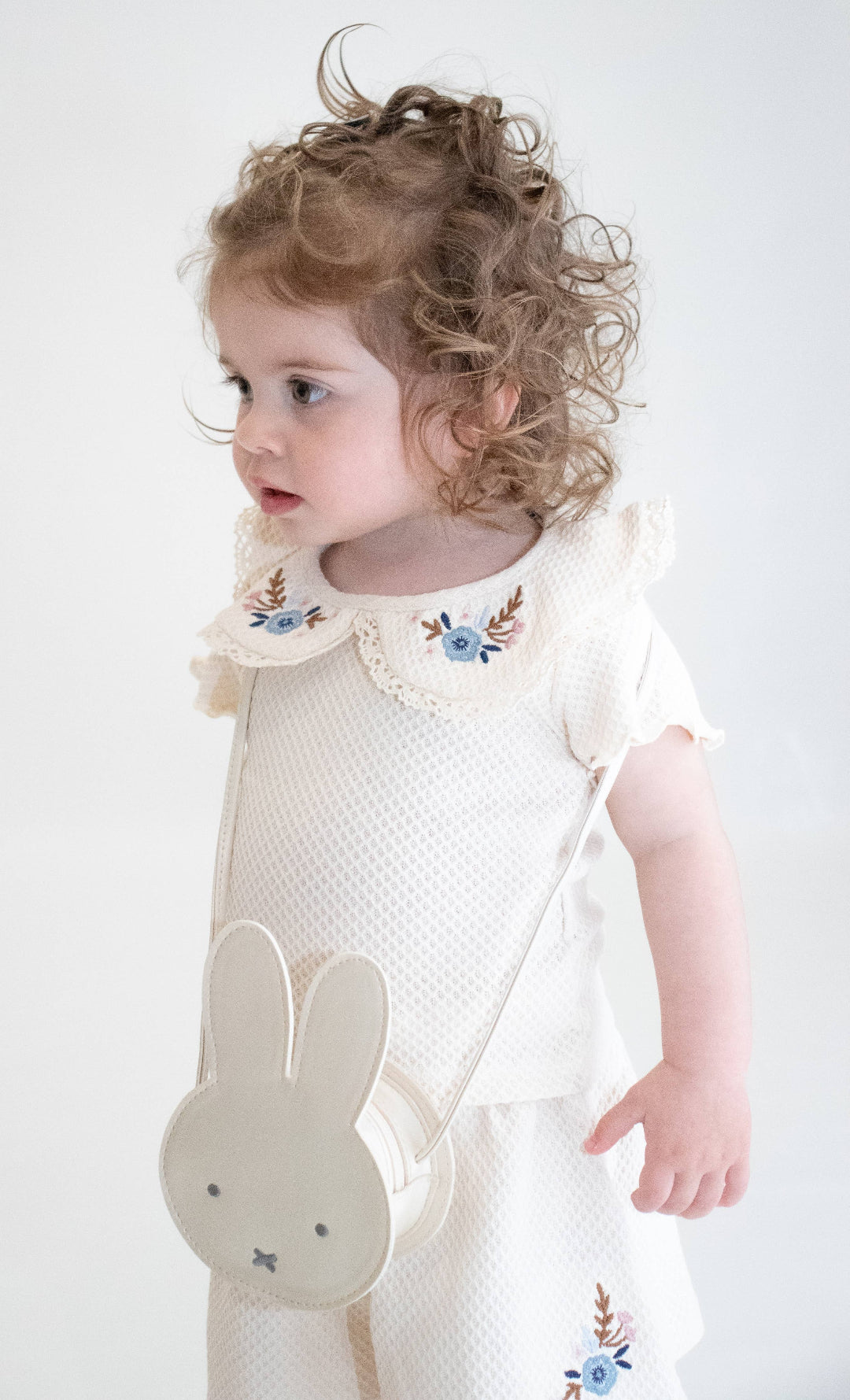 Child wearing a white outfit with floral details and a bunny bag on a plain background