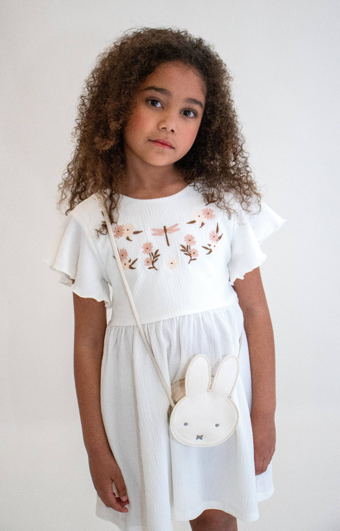 Young girl wearing a white dress with floral embroidery, holding a small white Miffy bag