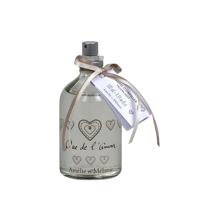 A clear glass bottle with a spray nozzle, containing a white liquid, with a label that reads 'Que de L'amour' and is adorned with heart designs and the brand name 'Amélie & Méléne'.
