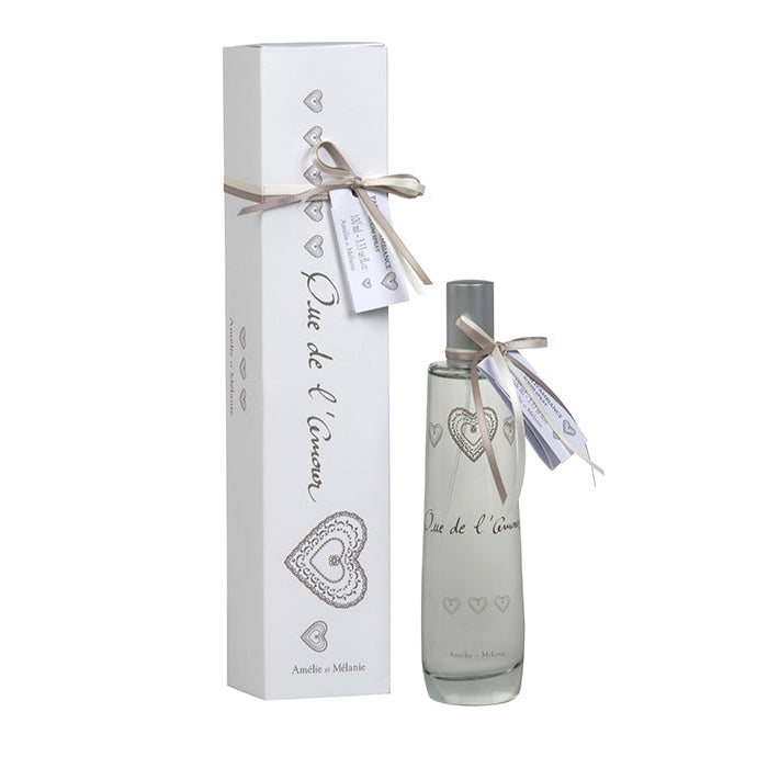 A room spray bottle with white label and decorative elements, tied with a string on top, presented next to its white packaging box adorned with hearts.