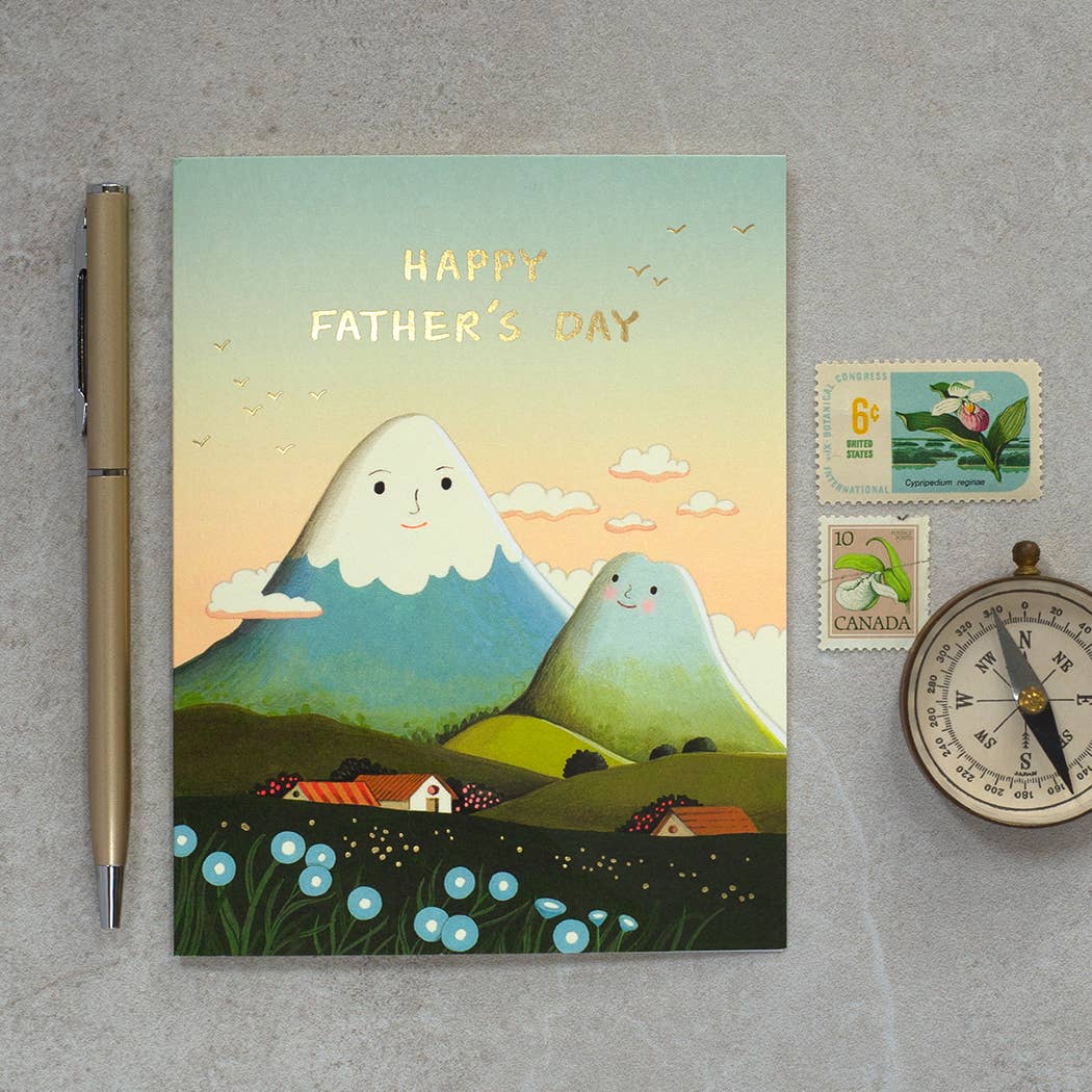 Father's Day card with cartoon mountains on a gray surface with a pen, stamps, and clock.
