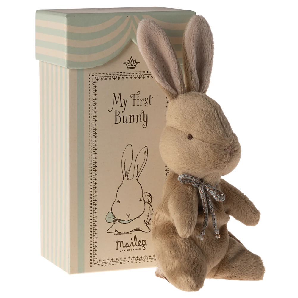 A plush toy rabbit with a brown body, beige ears, and a blue ribbon around its neck, sitting next to its beige and blue striped gift box with 'My First Bunny' text on it.
