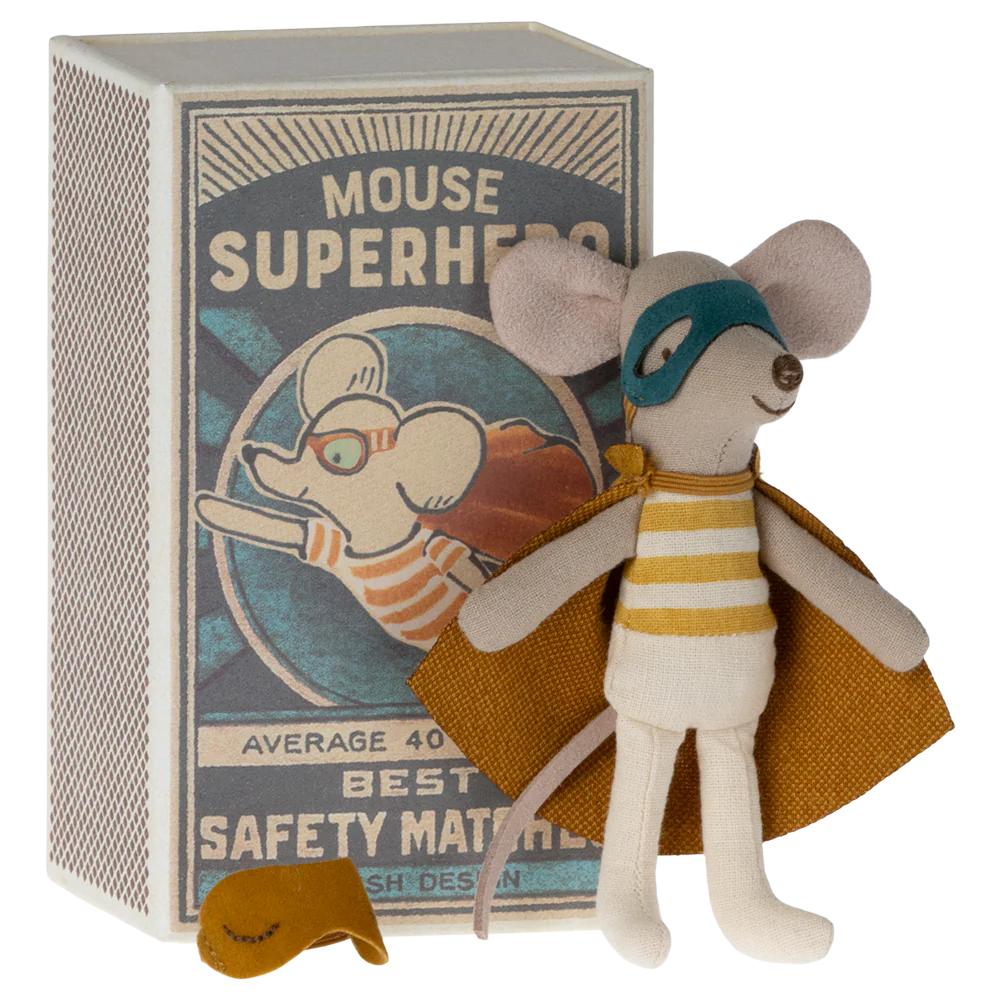 A toy mouse dressed as a superhero with a cape, standing next to a matchbox-sized cardboard box with a cartoon illustration.