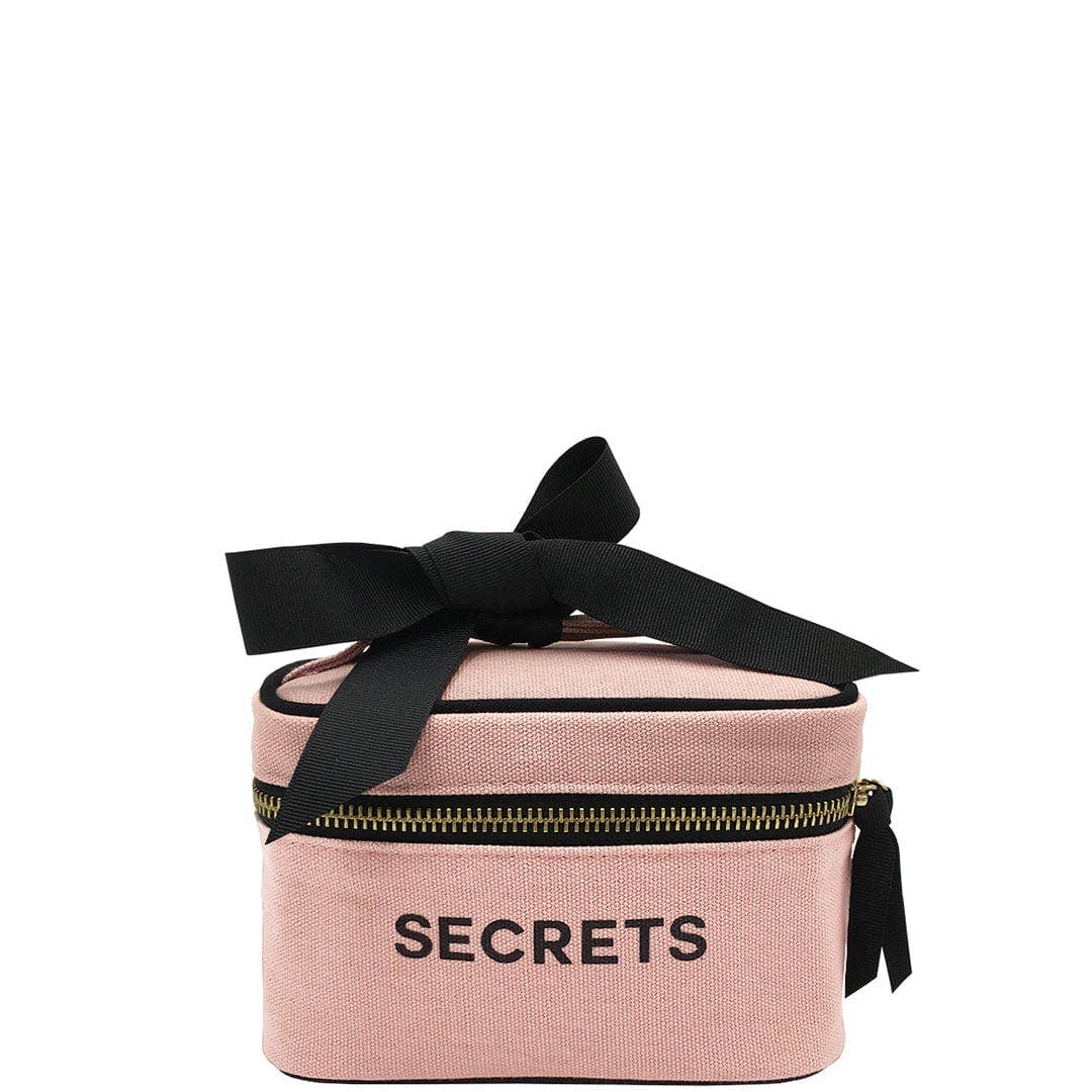 A rectangular blush pink mini beauty box with the word 'SECRETS' printed on the front, adorned with a black bow on top.