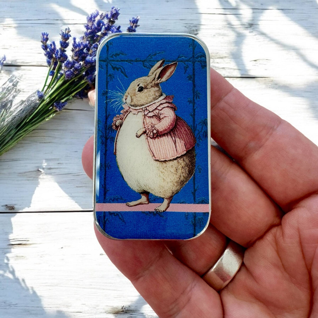 Hand holding a small rectangular tin with a rabbit illustration on a wooden surface with lavender.