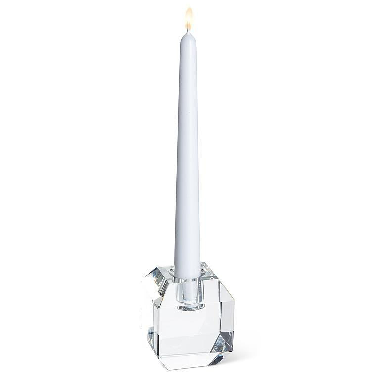 A white taper candle with a small flame, standing in a clear faceted glass base on a plain white background.