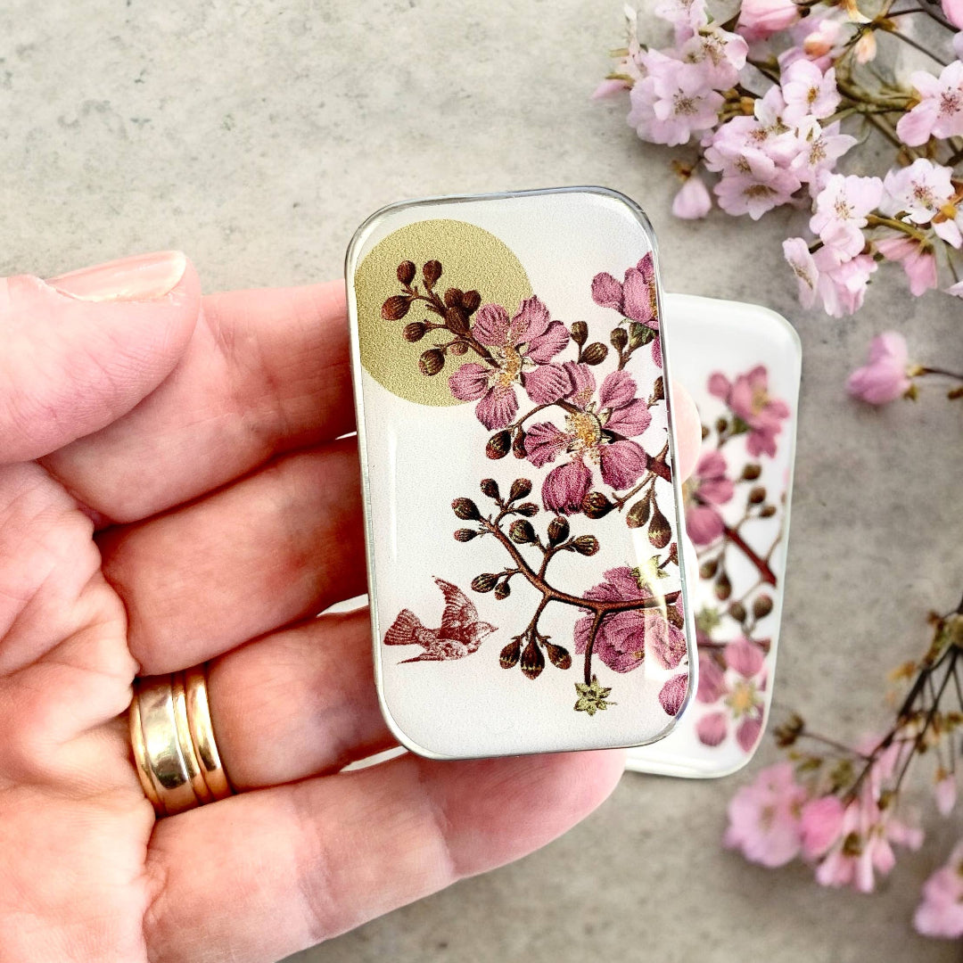 A small notions tin with a floral pattern, held in a hand, accompanied by real cherry blossoms in the background.