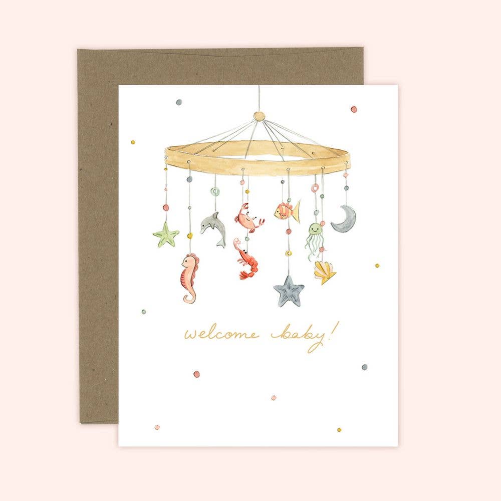 A 'Welcome Baby' greeting card featuring a watercolor illustration of a sea-themed mobile with animals and a moon, with a kraft envelope in the background.
