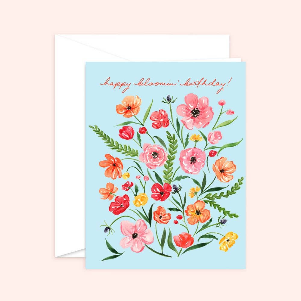 A floral birthday card with the text 'Happy Bloomin' Birthday!' featuring a watercolor illustration of blooming flowers in various colors on a light blue background. A white envelope is also shown.