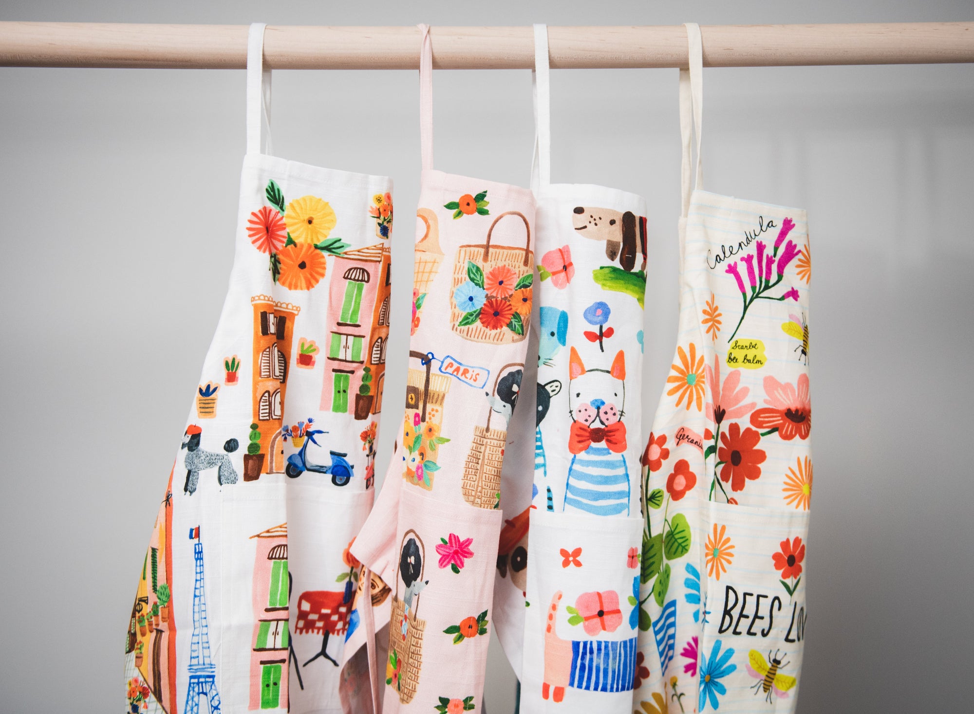 Four colorful aprons with various designs hanging on a rod against a gray background