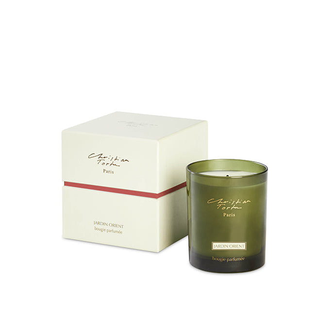 A green Christian Tortu scented candle with the label 'Jardin Orient' on the candle itself and a beige box with the brand and product name in the background.