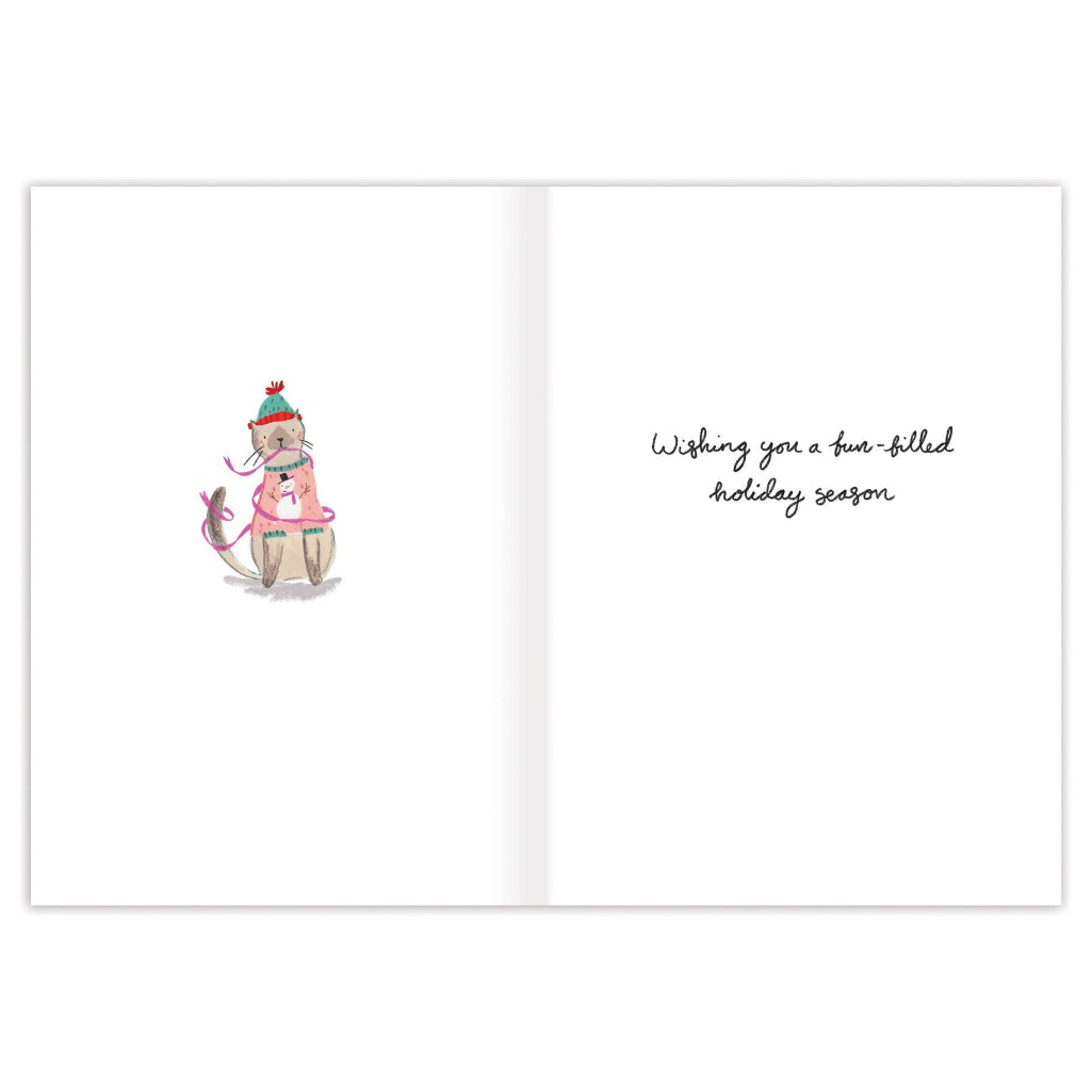 An open greeting card with a white background. The left side features a small watercolor illustration of a cat wearing a red and green striped knit hat and a pink sweater, sitting 