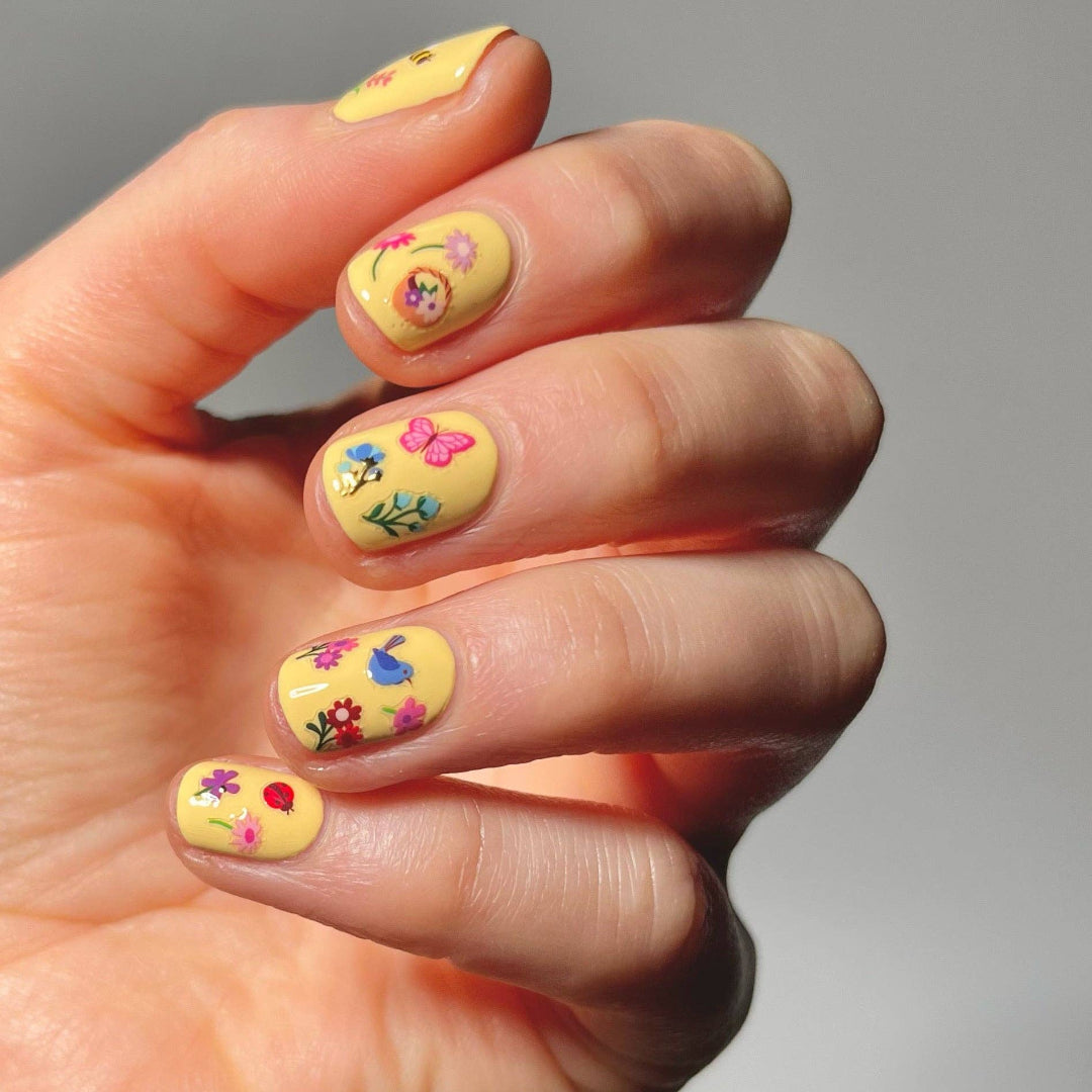 Hand with yellow nail polish featuring floral and butterfly nail stickers on a neutral background