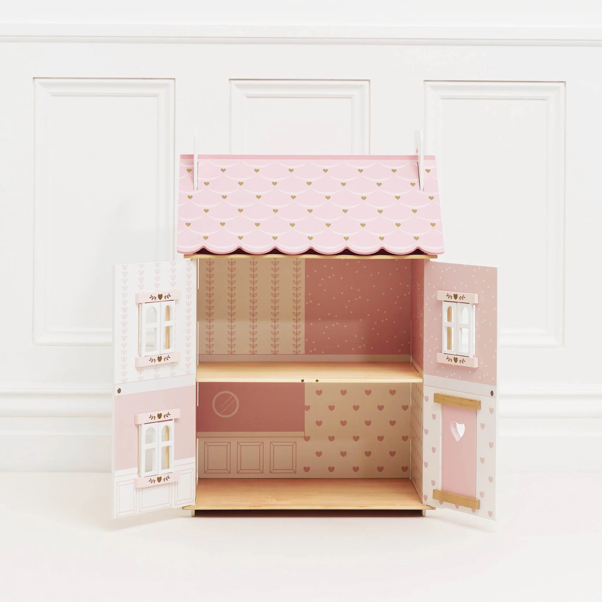A pink and white wooden dolls house with a scalloped roof, opening doors, and shutters. It has a natural wood look on the floors and decorative heart motifs.