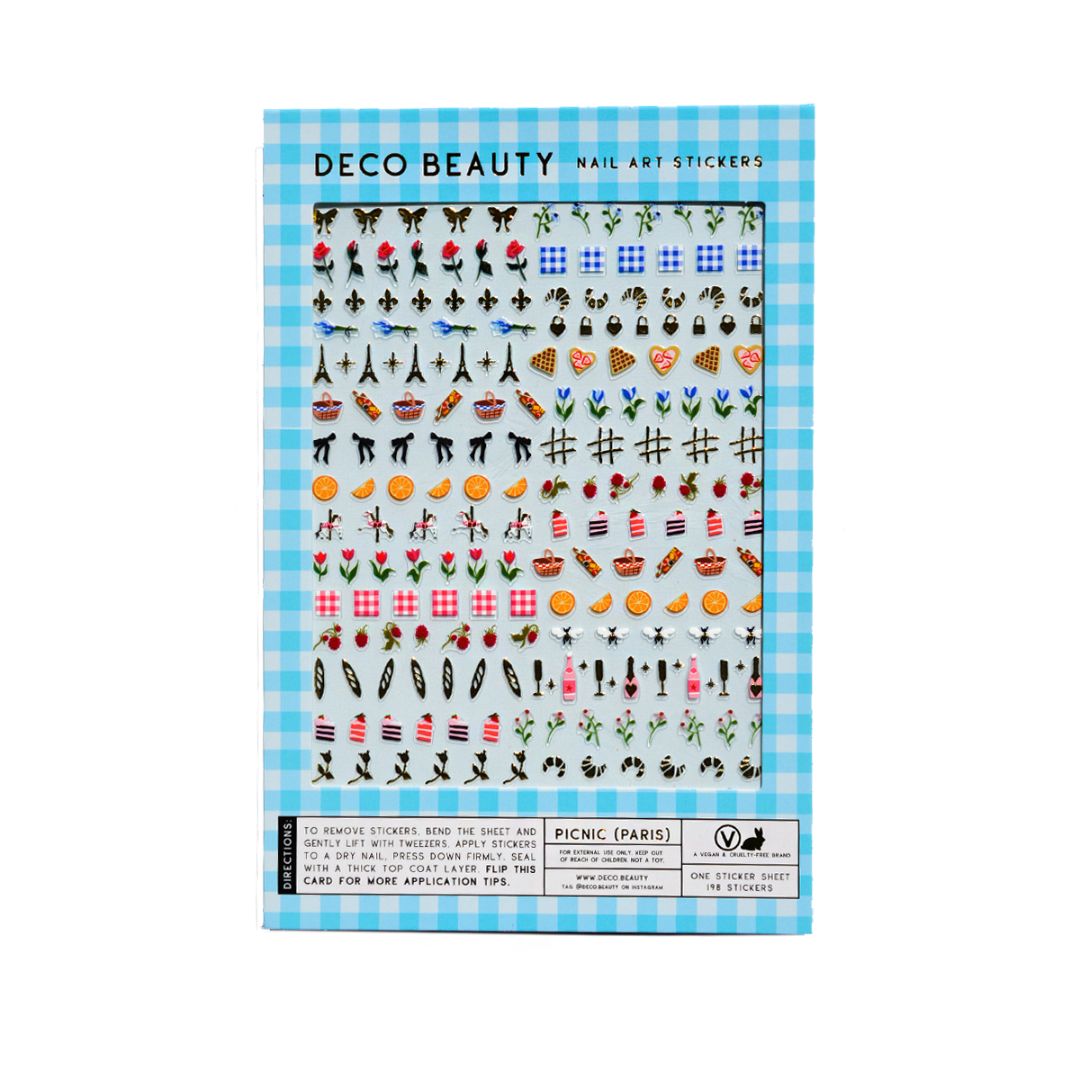 A sheet of Deco Beauty nail art stickers featuring a light blue gingham background and rows of colorful decorative icons, including flowers, hearts, Eiffel Towers, umbrellas, and food motifs.