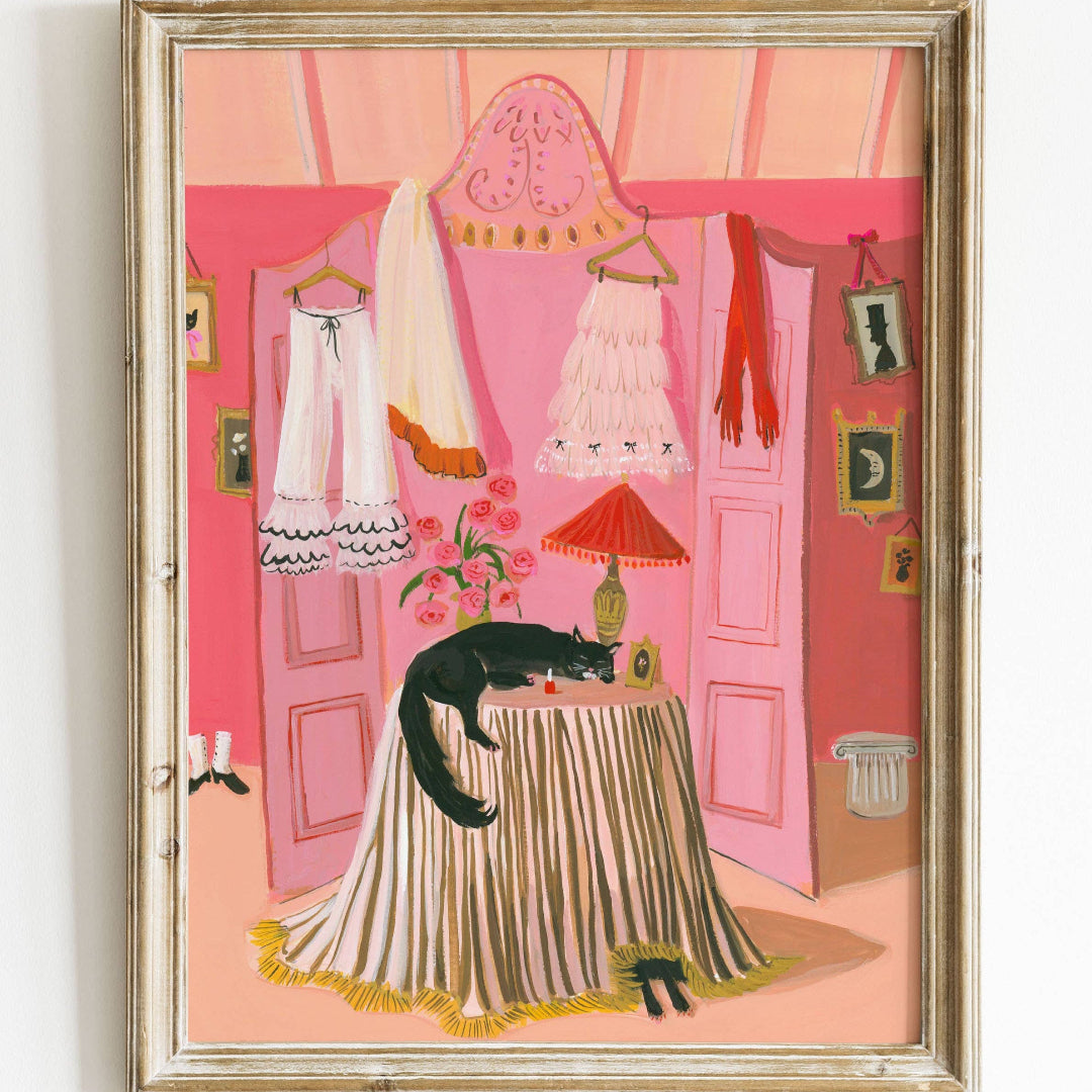 A framed art print featuring a vibrant illustration of a cat sitting on a dress, with a pink background and various decorative items in a room.