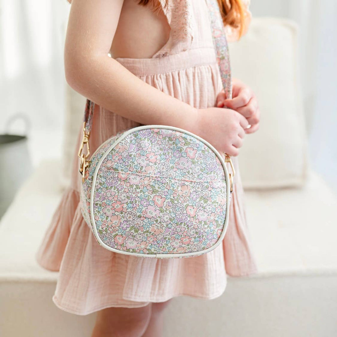 A child in a pink dress is holding a multicolored floral crossbody messenger bag with gold hardware.