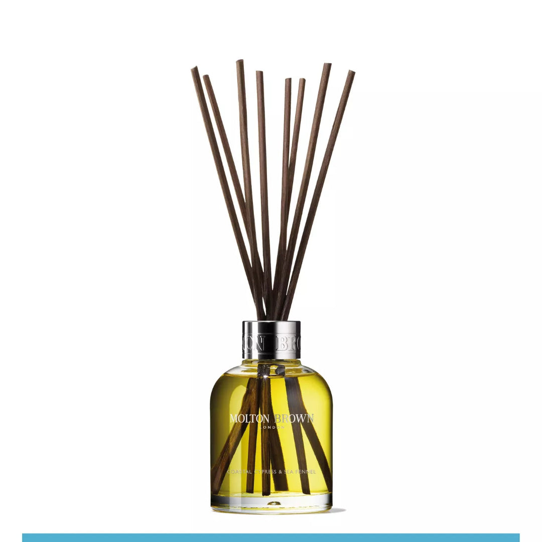 A Molton Brown aroma diffuser with several brown reeds inserted into a clear glass bottle filled with yellow liquid, set against a white background.